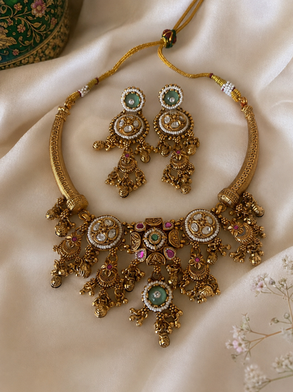 Rithwika traditional jewelry set displaying an antique gold-finished Hasli rigid collar necklace featuring round crystal polki medallions, a central cascading fringe with filigree gold drops, pink accent stones, and mint green emeralds, laid flat on soft cream satin fabric alongside matching long drop earrings.