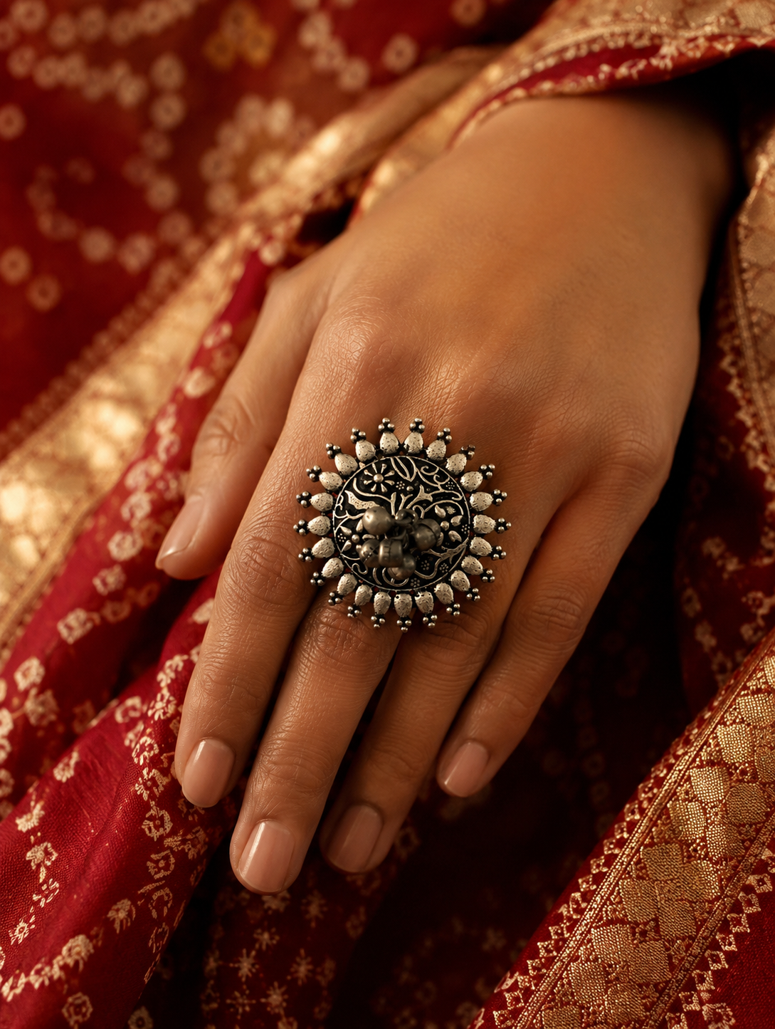 A close-up of a woman's hand resting on a red and gold traditional textile, showcasing a large, round oxidized silver statement ring with a pointed sunburst frame and a detailed floral engraved center.