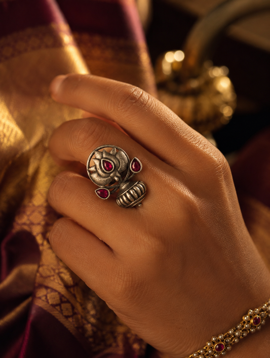 A close-up view of a woman's hand wearing an oxidized silver ring shaped like a lotus blossom with ruby-pink teardrop stones, set against a rich purple and gold silk saree.
