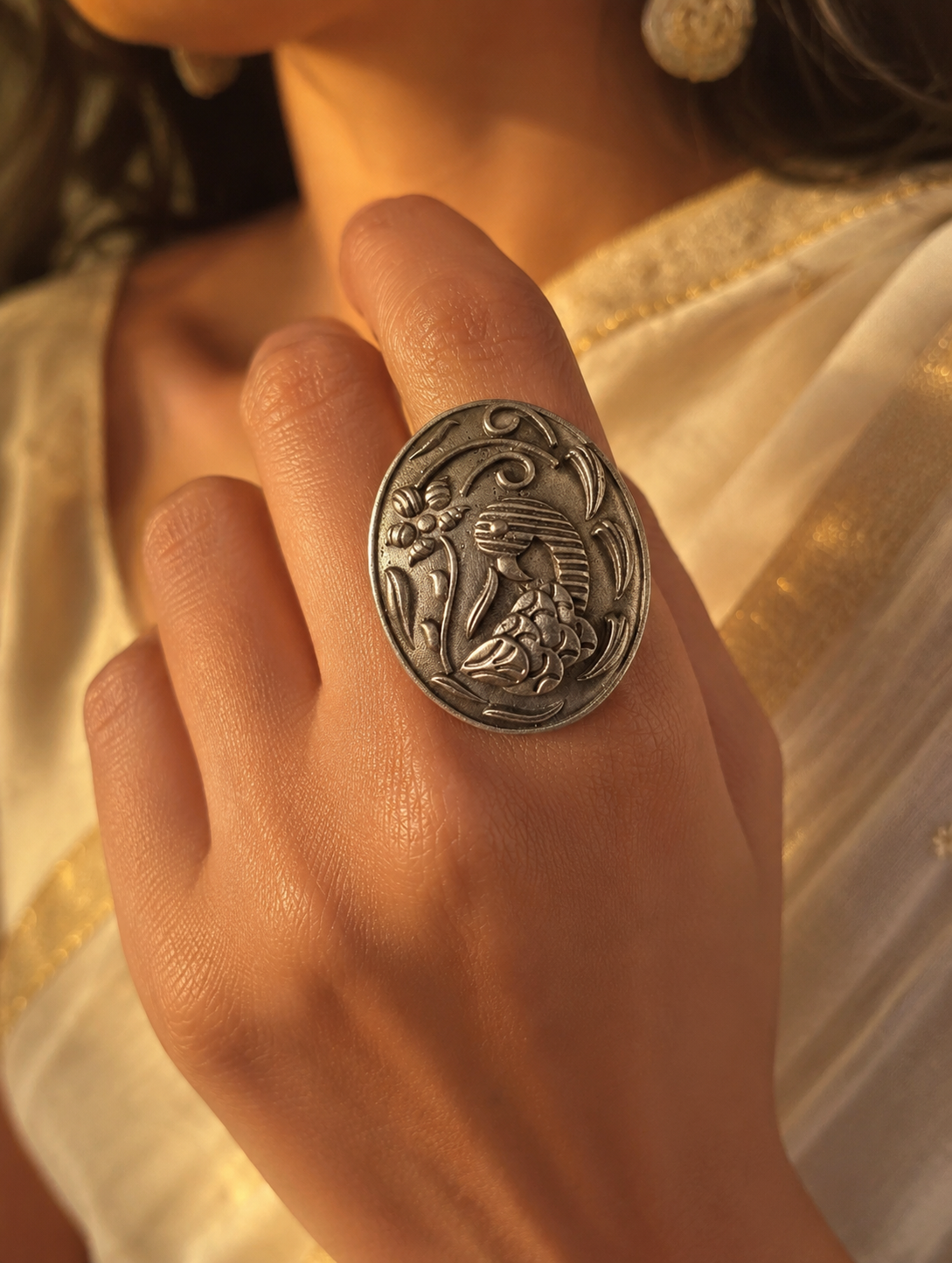 A close-up of a woman's hand showcasing a large, oval-shaped oxidized silver statement ring with an embossed traditional bird and floral design, paired with an elegant cream and gold saree.
