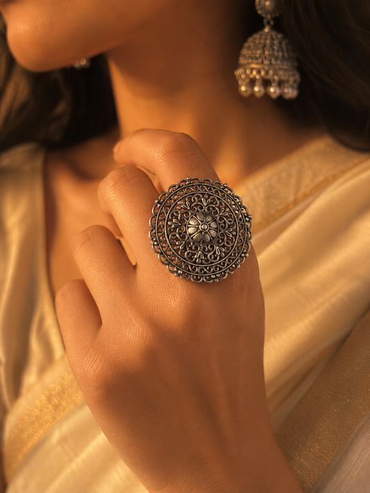 A close-up view of a woman's hand showcasing a large, round oxidized silver statement ring with intricate filigree patterns and a central flower motif, paired with a matching jhumka earring and a cream saree.