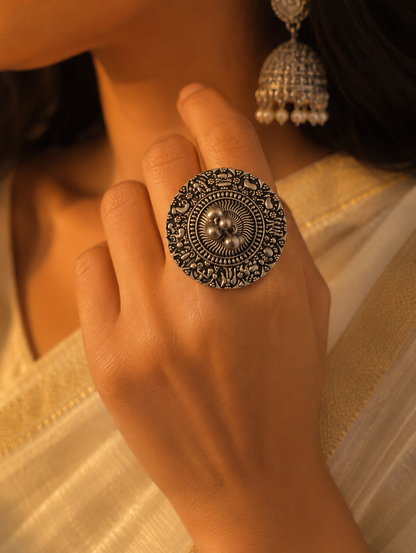 A close-up view of a woman's hand wearing a large, round oxidized silver statement ring with micro-engraved ethnic patterns and a center bunch of silver metallic beads, complemented by a cream saree and jhumka earring.
