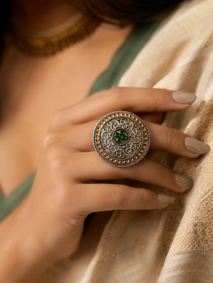 A close-up view of a woman's hand displaying a large, circular statement ring crafted in dual-tone oxidized silver and gold filigree, featuring a small cluster of four green stones in the center, styled with an off-white and gold traditional outfit.