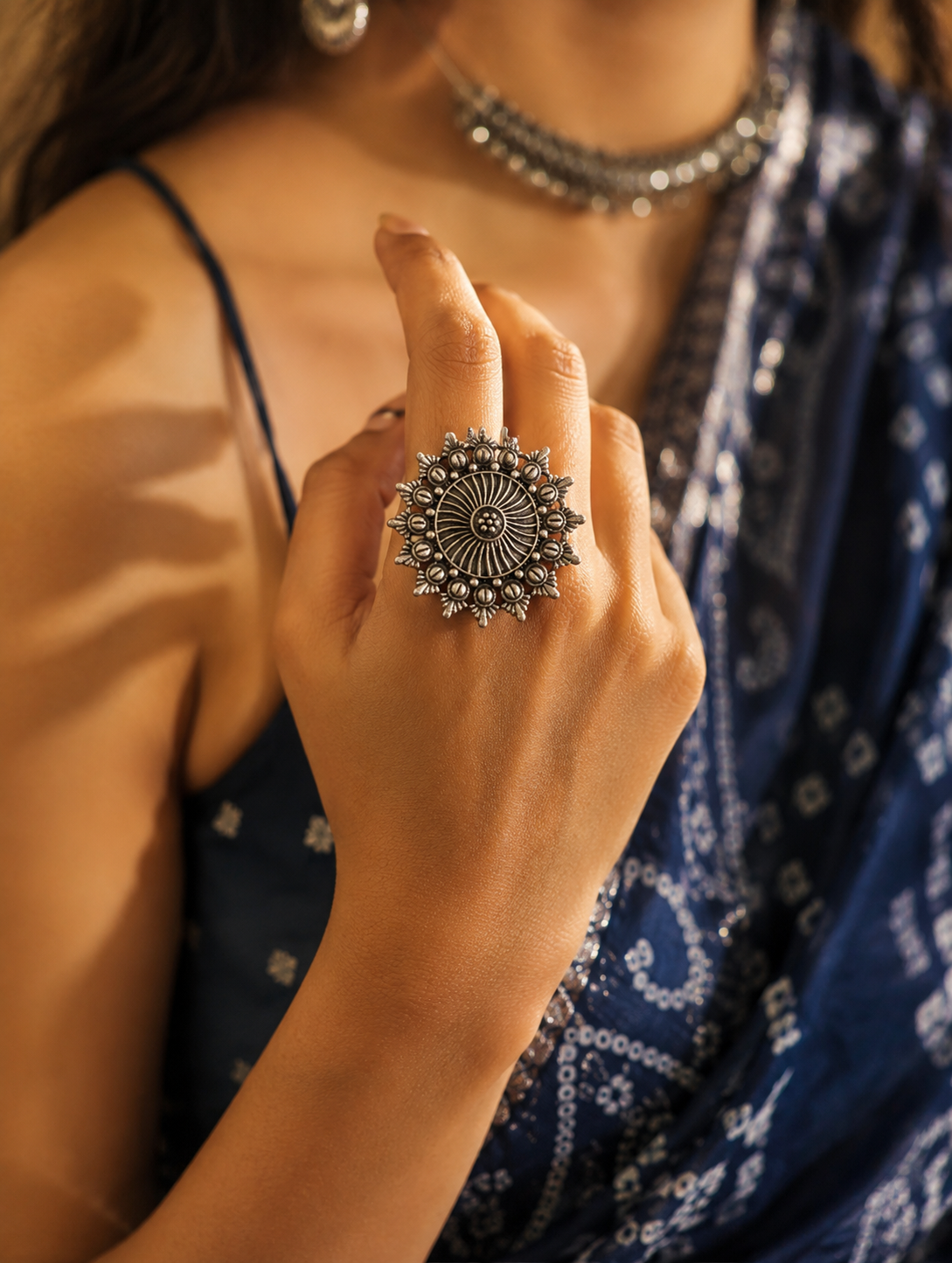 A close-up view of a woman's hand showcasing a large, round oxidized silver statement ring with a pointed sunburst edge and a spiraled, wheel-like pattern at its center, paired with a dark blue traditional saree and matching silver necklace.