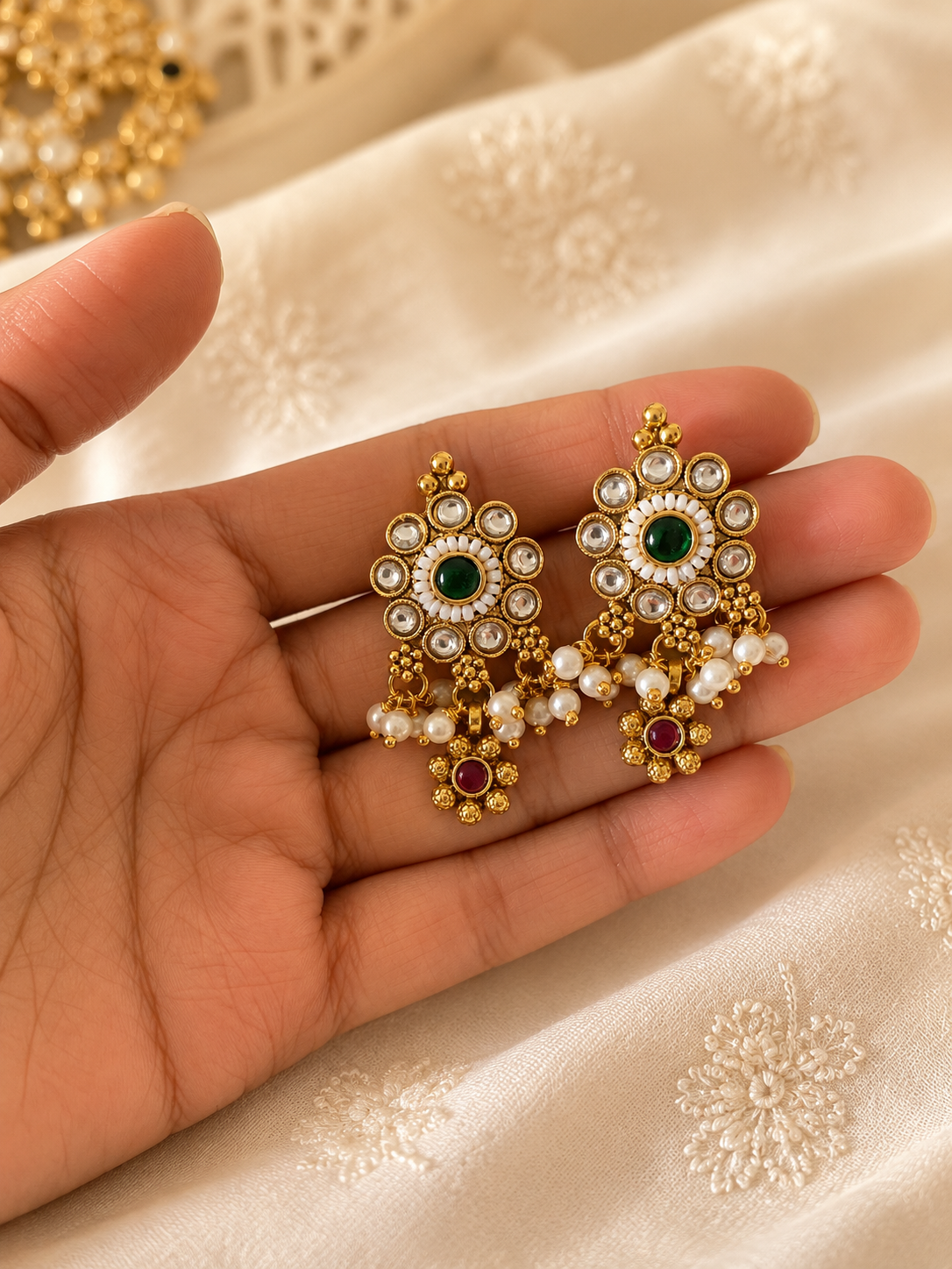 Gold earrings with green and red stones held in a hand on a beige fabric background