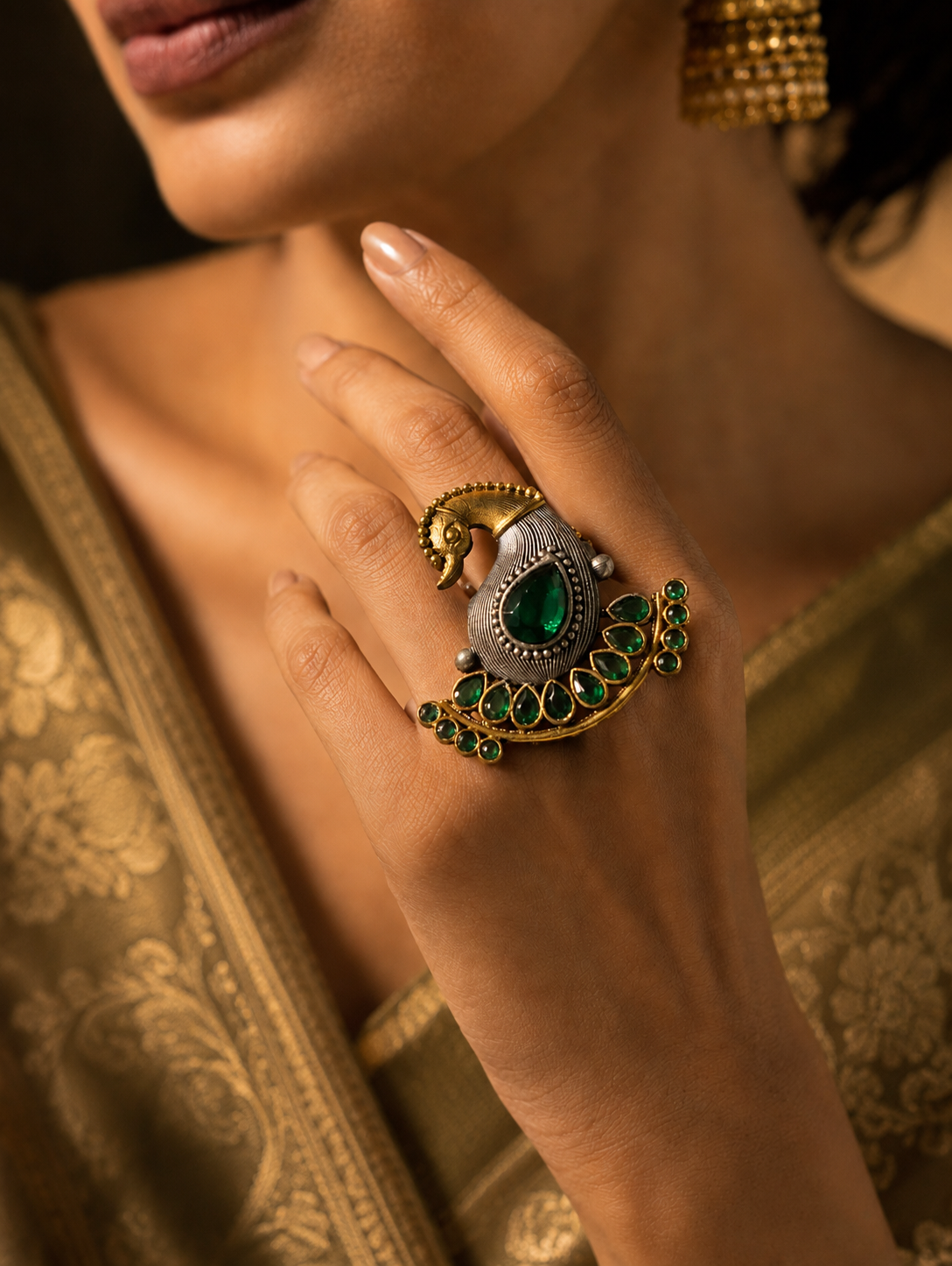 A close-up view of a woman's hand showcasing an ornate dual-tone peacock statement ring. The ring features a gold-toned bird head and a textured oxidized silver body, beautifully accented by a fan of royal blue teardrop gemstones, styled alongside a gold silk saree.