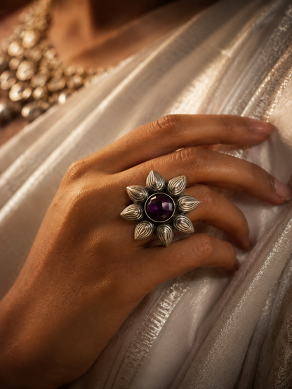 A close-up view of a woman's hand showcasing a large silver statement ring shaped like a blooming flower with textured petals, featuring a prominent round purple gemstone at its center, set against a shimmering silver traditional fabric.