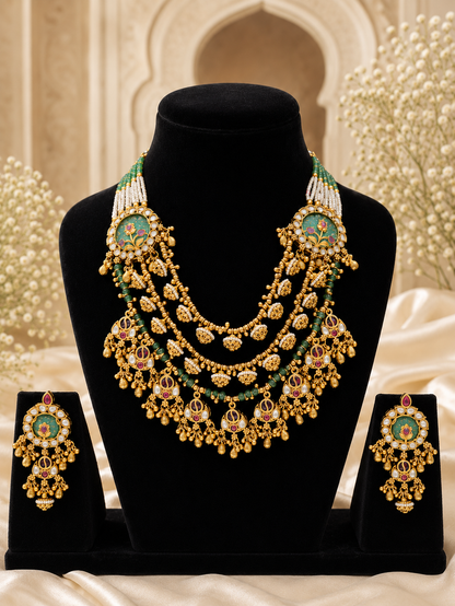 An elegant antique gold-plated multi-layered bridal necklace set with intricate green enamel work, ruby accents, pearls, and matching chandelier earrings, displayed on a black bust against a soft, festive background.