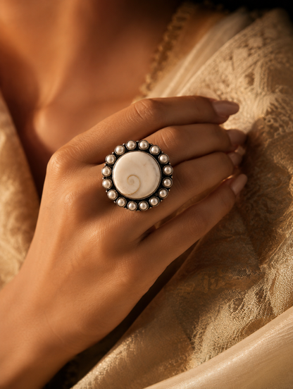 A close-up view of a woman's hand showcasing a large, round oxidized silver statement ring. The ring features a polished white shankha shell center with a natural spiral pattern, surrounded by a delicate border of small white pearls, set against a gold silk garment.