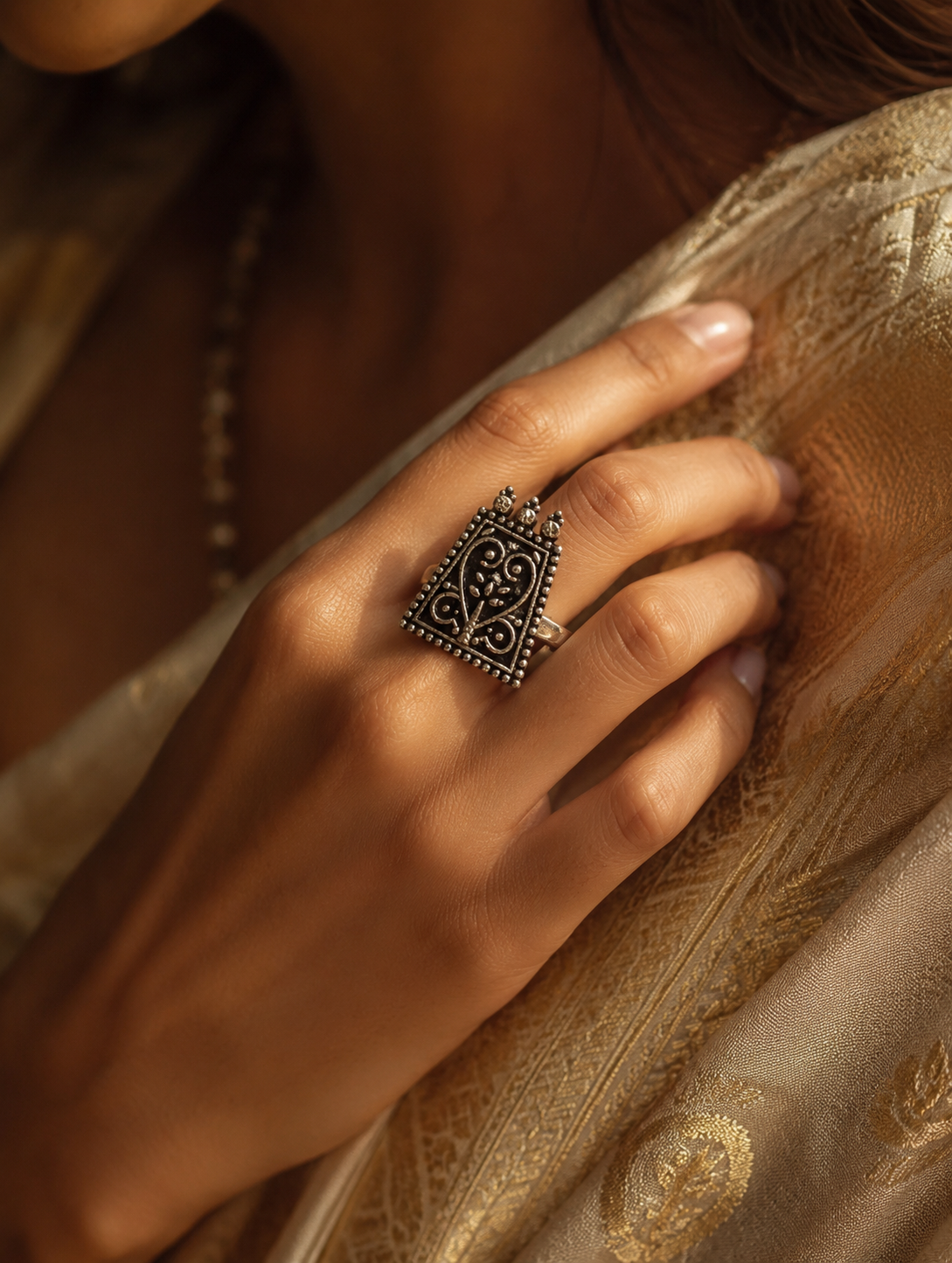 A close-up view of a woman's hand showcasing a trapezoid shield-shaped oxidized silver ring with intricate filigree heart and scroll motifs, styled against a gold embroidered traditional saree.