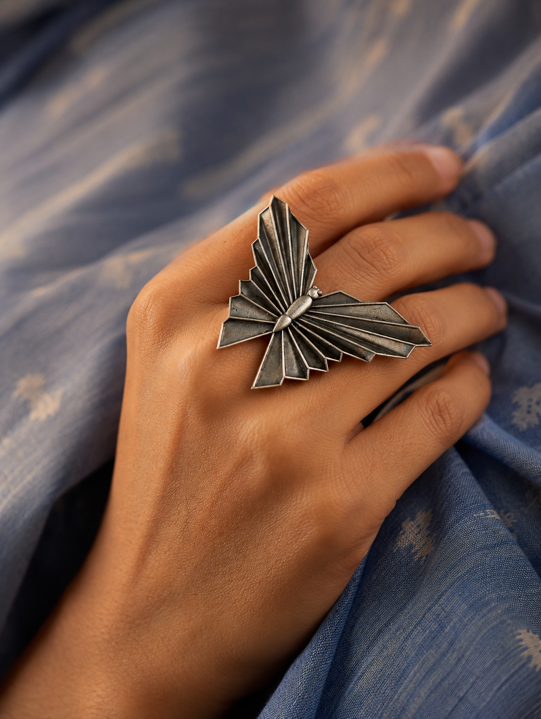 A close-up view of a woman's hand showcasing an oversized oxidized silver statement ring sculpted in the shape of a butterfly with modern, geometric pleated wings, set against a soft blue textured fabric.