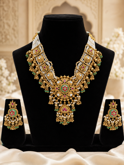 An intricate antique gold bridal necklace and matching earrings set from the RajRatna collection, displayed on black velvet stands. The jewelry features detailed Kundan craftsmanship, a central green enameled floral motif with ruby accents, multi-strand white seed pearl bands, and dangling gold and emerald green bead clusters.