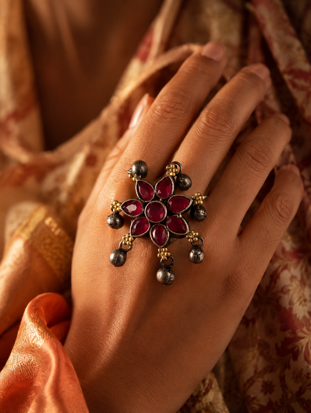 A close-up view of a woman's hand displaying a unique oxidized silver statement ring designed as a blooming flower with teardrop-shaped ruby-red stones, accented by small gold beads and playful dangling silver ghungroo charms, set against a peach and beige ethnic outfit.