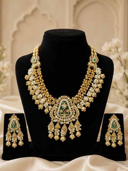 An elegant traditional Indian bridal jewelry set from the YuvRaani collection displayed on black velvet busts. The set includes a heavy gold-polished necklace and matching tiered dangle earrings, detailed with intricate gold beadwork, clusters of small white pearls, and a prominent green enameled central pendant accented with small pink rubies and clear stones.