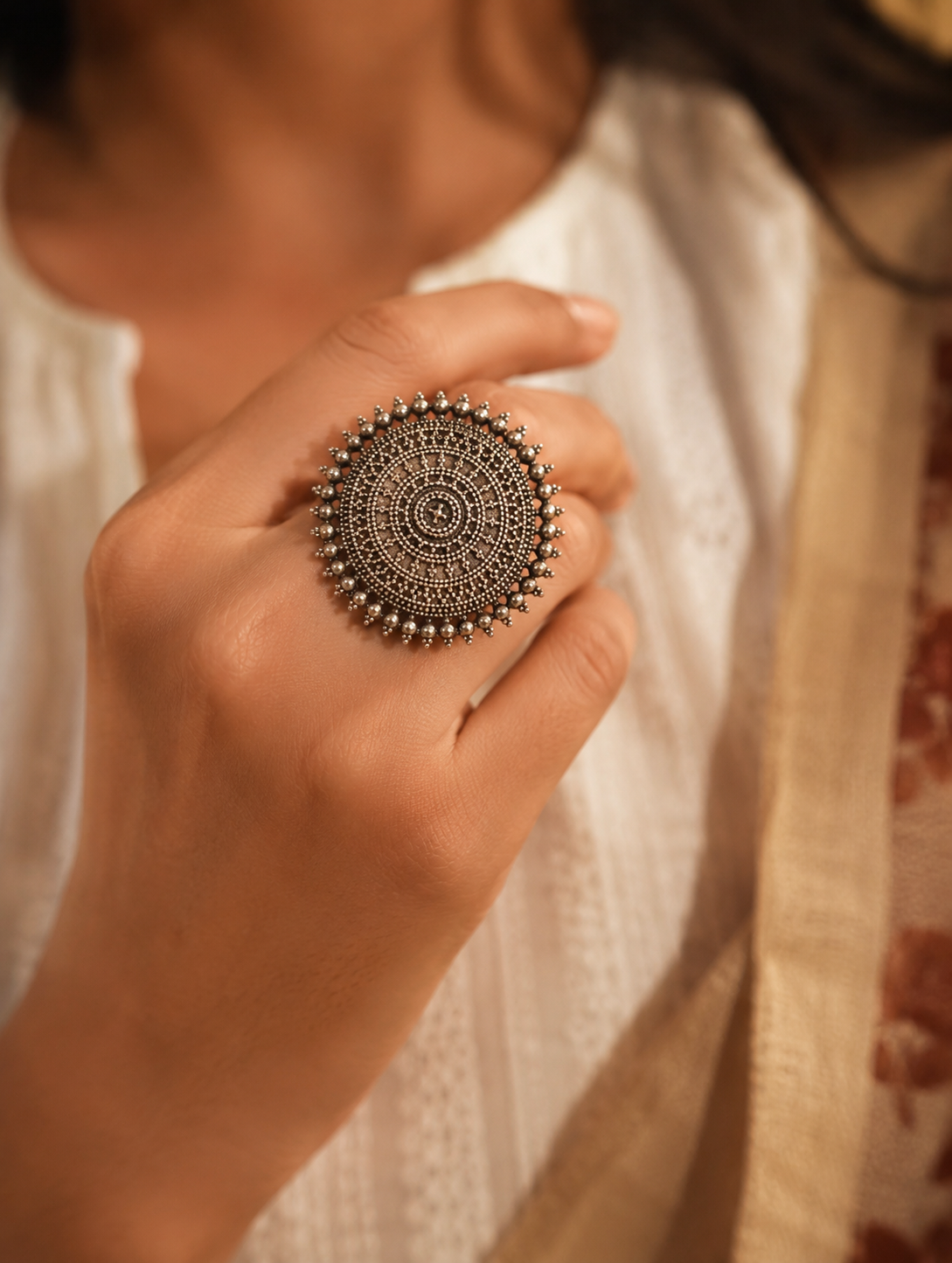 A close-up view of a woman's hand displaying a large, circular oxidized silver statement ring with highly detailed concentric patterns reminiscent of a mandala, finished with a spiked metallic bead border and paired with a white textured top.