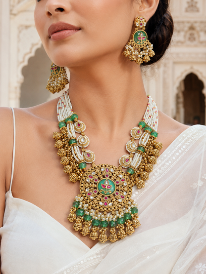Woman wearing a gold necklace and earrings with green and red accents in an outdoor setting.