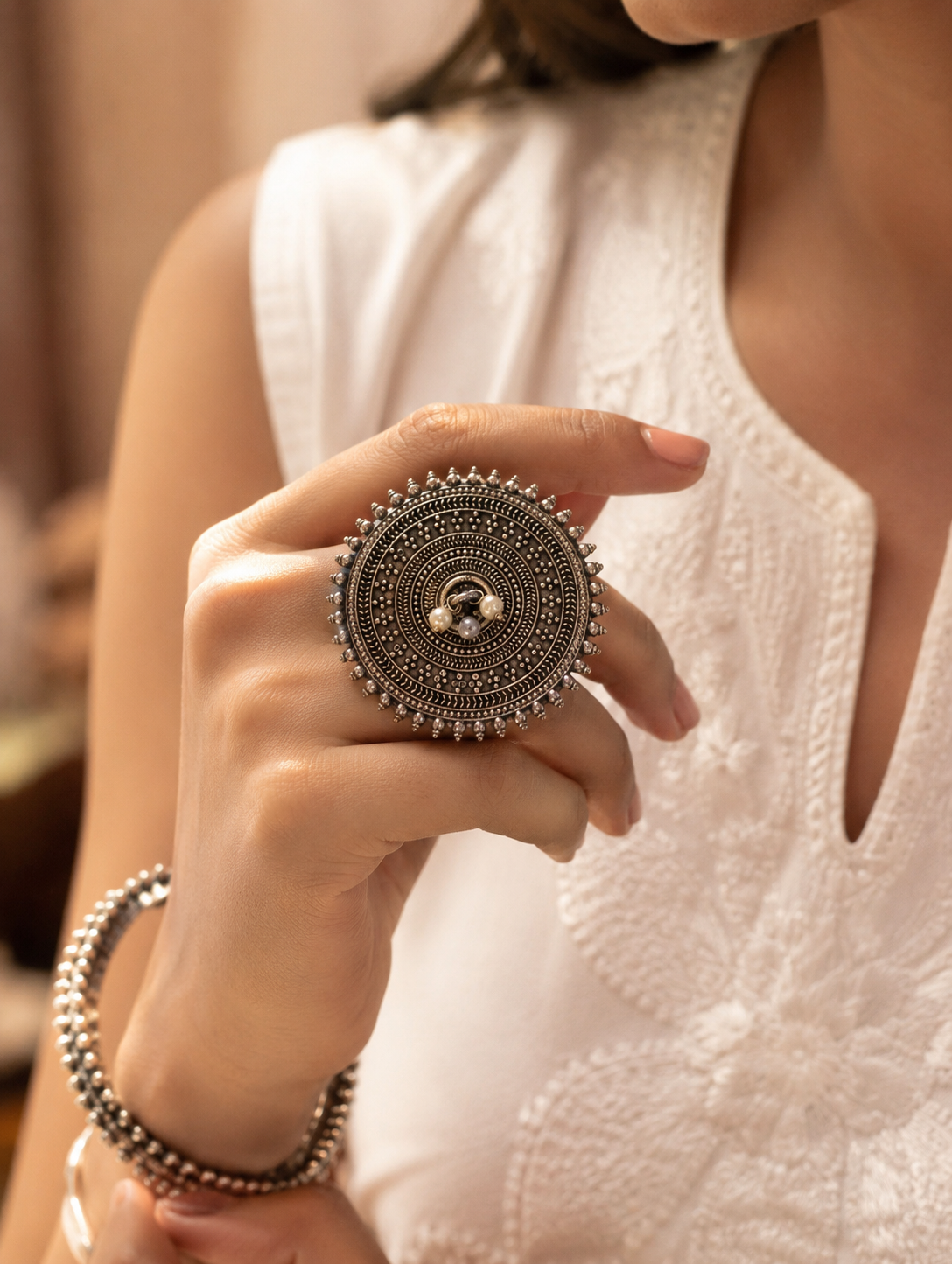 A close-up view of a woman's hand showcasing an oversized, round oxidized silver statement ring detailed with concentric geometric motifs, a spiked rava border, and a cluster of tiny pearls in the absolute center, styled with a white embroidered ethnic outfit.