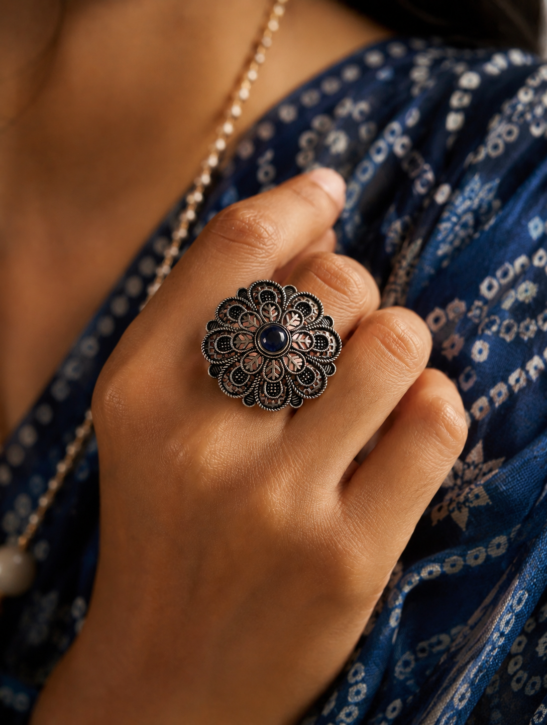 A close-up view of a woman's hand displaying a large, circular oxidized silver statement ring designed with a detailed, scalloped floral mandala pattern and a small, round dark blue gemstone at its center, styled against a blue bandhani print saree.
