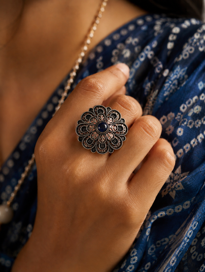 A close-up view of a woman's hand displaying a large, circular oxidized silver statement ring designed with a detailed, scalloped floral mandala pattern and a small, round dark blue gemstone at its center, styled against a blue bandhani print saree.