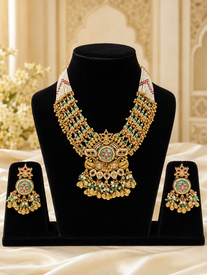 Traditional Indian antique gold bridal necklace and matching earrings set displayed on black busts. The intricate necklace features multi-strand white seed pearls, multi-layered gold beads, and a large central pendant adorned with green and pink Meenakari enamel, kundan stones, and dangling gold and green bead clusters.