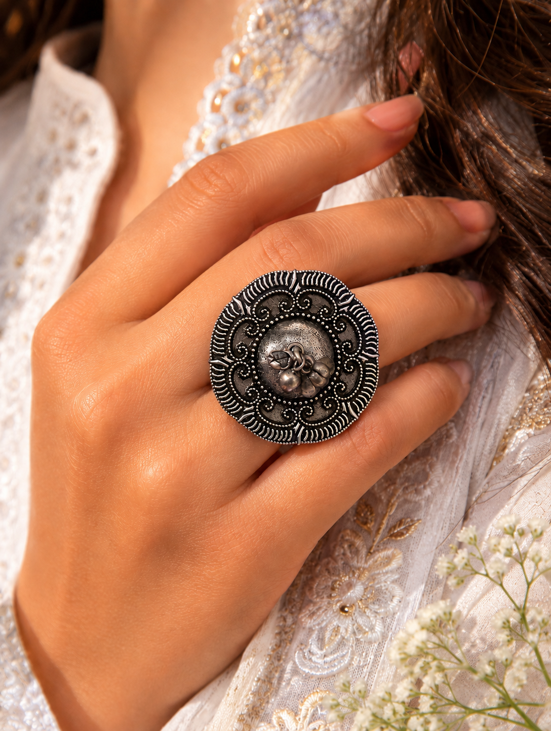 A close-up view of a woman's hand showcasing an oversized circular statement ring in oxidized silver. The ring features a raised textured dome center with a pair of tiny dangling ghungroo charms, encircled by a wide, detailed border of intricate metal scrollwork and a dark, contrasting radial edge, styled with a white and gold embroidered outfit.