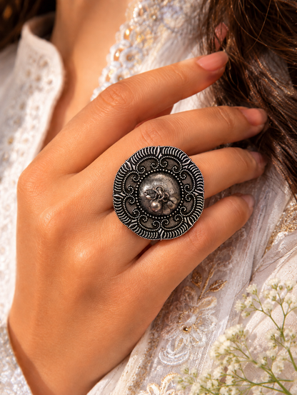 A close-up view of a woman's hand showcasing an oversized circular statement ring in oxidized silver. The ring features a raised textured dome center with a pair of tiny dangling ghungroo charms, encircled by a wide, detailed border of intricate metal scrollwork and a dark, contrasting radial edge, styled with a white and gold embroidered outfit.