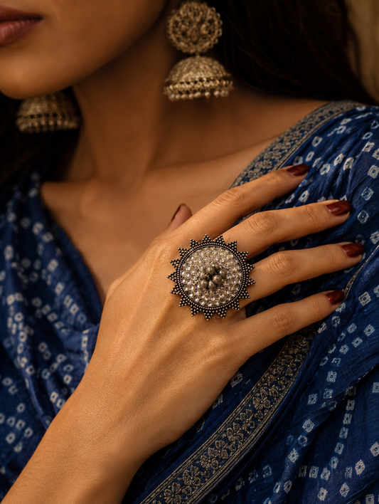 A close-up view of a woman's hand showcasing a large, round oxidized silver statement ring studded with a cluster of clear crystals or miniature mirror stones, finished with a spiked sunburst metallic border, styled with matching jhumka earrings and a blue bandhani print saree.