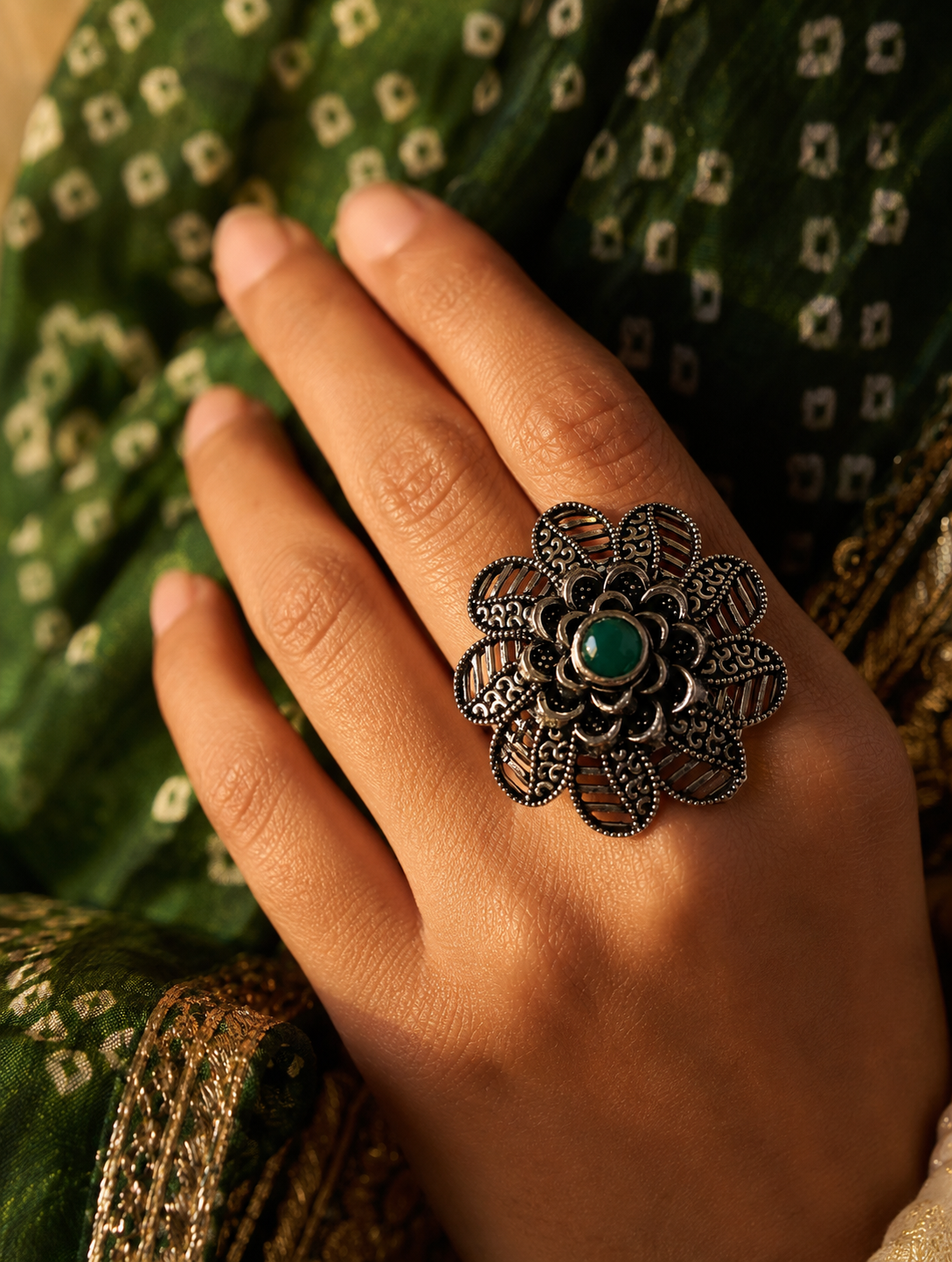 A close-up view of a woman's hand displaying a large, circular oxidized silver statement ring designed with a detailed, scalloped floral mandala pattern and a small, round green gemstone at its center, styled against a green bandhani print fabric.