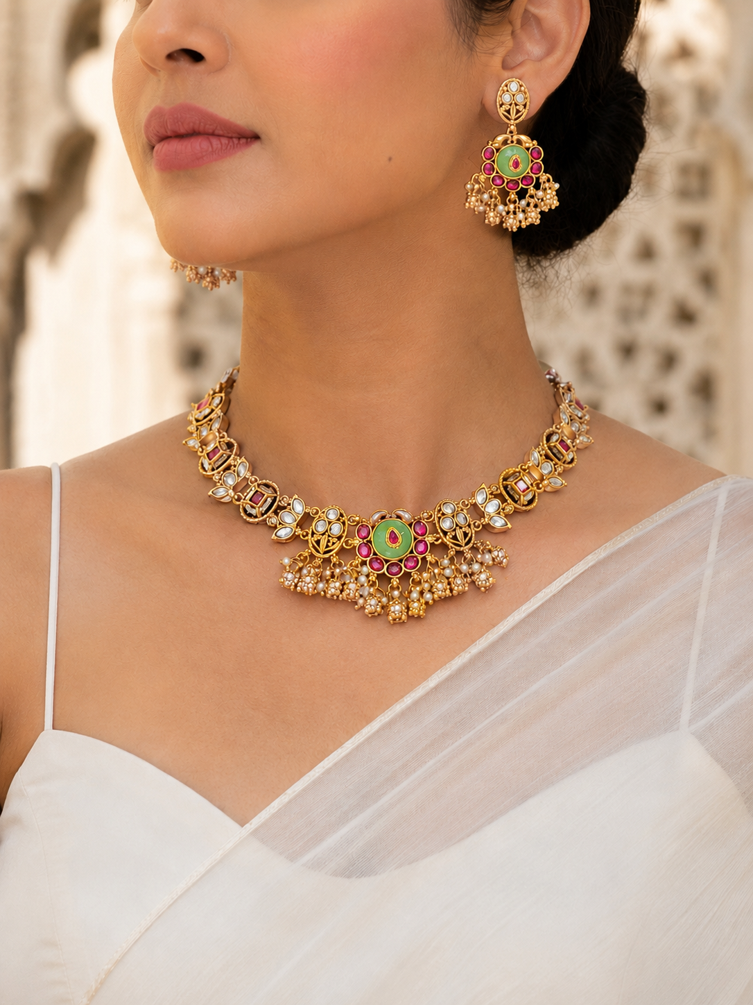 Close-up of a model wearing a traditional Indian antique gold choker necklace and matching round drop earrings, paired with a white saree. The intricate necklace features white kundan-style stones and a central floral motif with a light mint-green enamel center, pink stone accents, and delicate hanging gold beads and pearls.