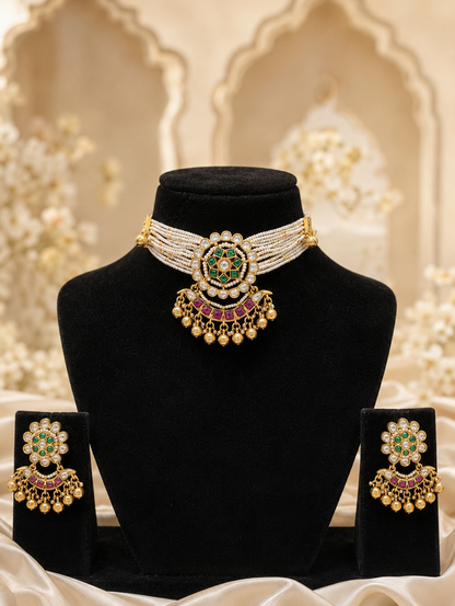 Heritage jewelry set featuring a wide multi-strand white seed pearl choker necklace supporting a round gold medallion pendant with a star-shaped green emerald center, ruby accents, and a dangling fringe of gold beads, showcased on a black velvet stand alongside matching floral drop earrings.
