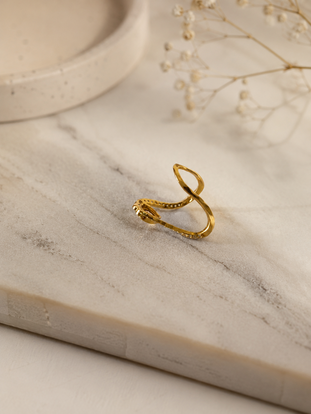 Side profile perspective of a gold-toned V-shaped Vanki ring laying on a white marble surface, showing the open curved band structure and stone setting details.