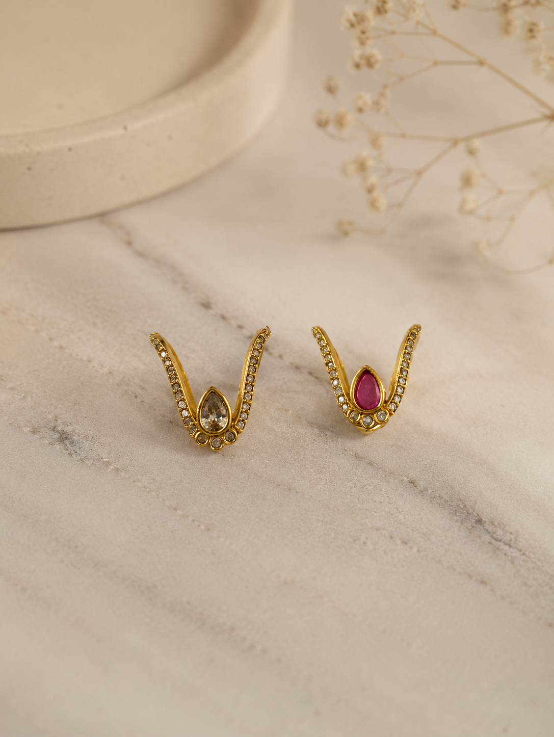 A pair of gold-toned V-shaped Vanki rings displayed side by side on marble, each featuring pave crystal-paved arms with a center teardrop stone, one in champagne white crystal and the other in ruby pink.