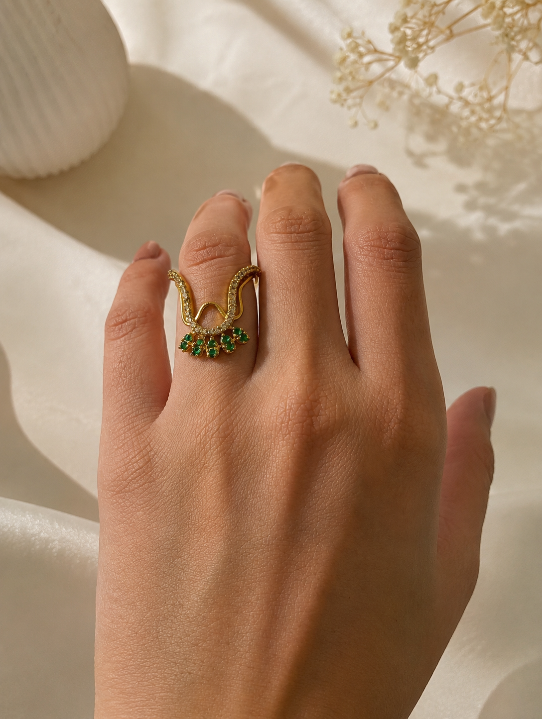 Close-up of a model's hand wearing a gold-toned Vanki ring on her index finger, displaying the V-shaped chevron curve with pave crystal accents and an emerald-green stone fan fringe.