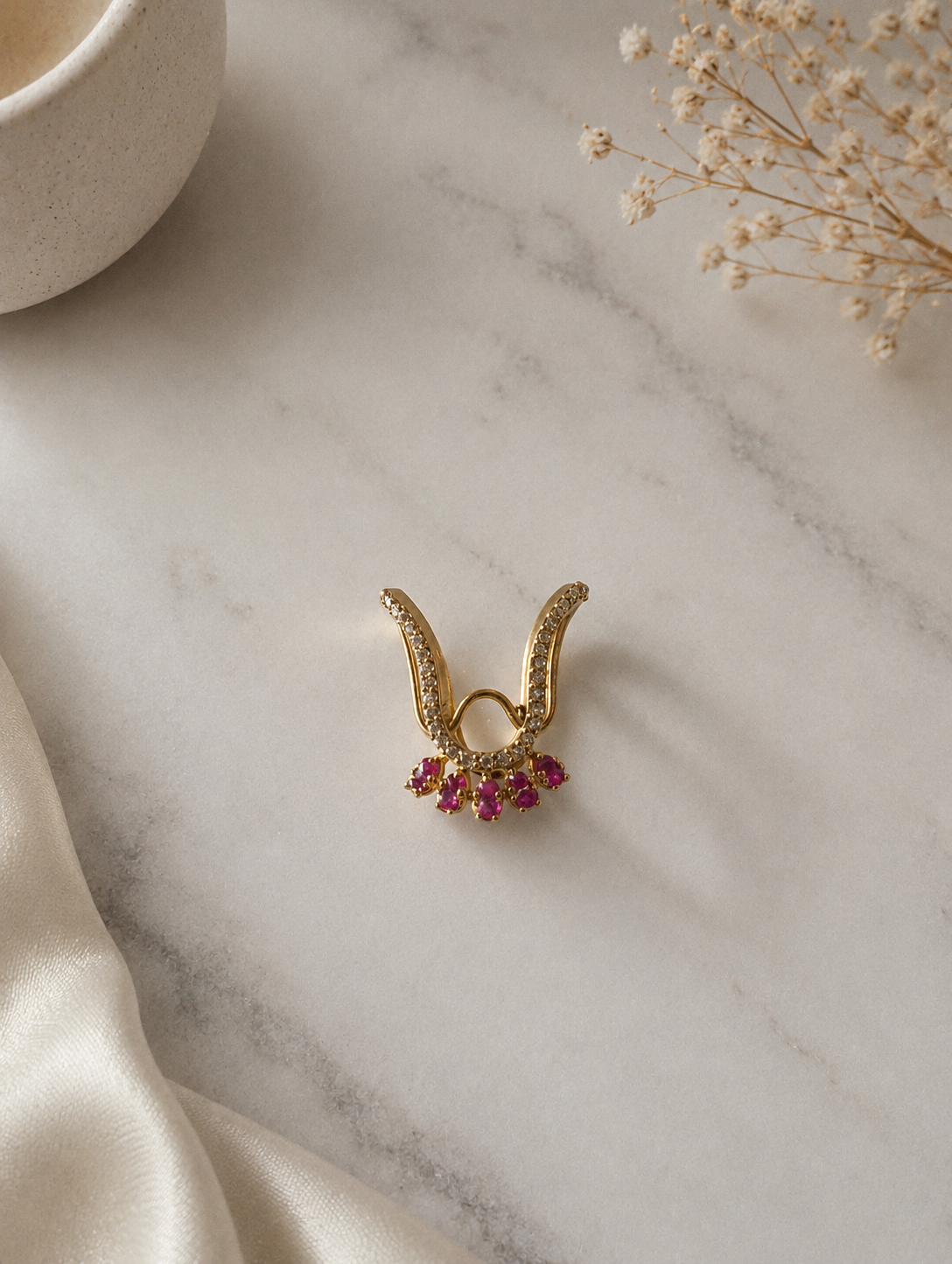 A gold-toned Vanki chevron ring with pave cz-lined arms and a fan fringe of five vibrant ruby-pink stones at the apex, placed on a beige marble surface next to white satin.