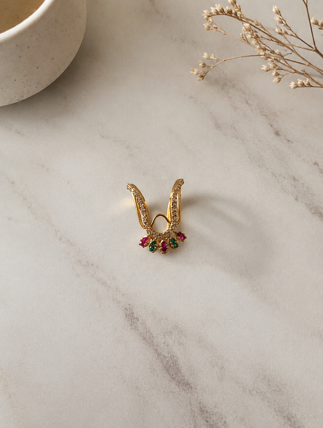 A gold-toned V-shaped Vanki ring featuring pave CZ crystals along the chevron band and a fan-shaped fringe of alternating ruby-red and emerald-green stones at the bottom tip, displayed on a marble surface.