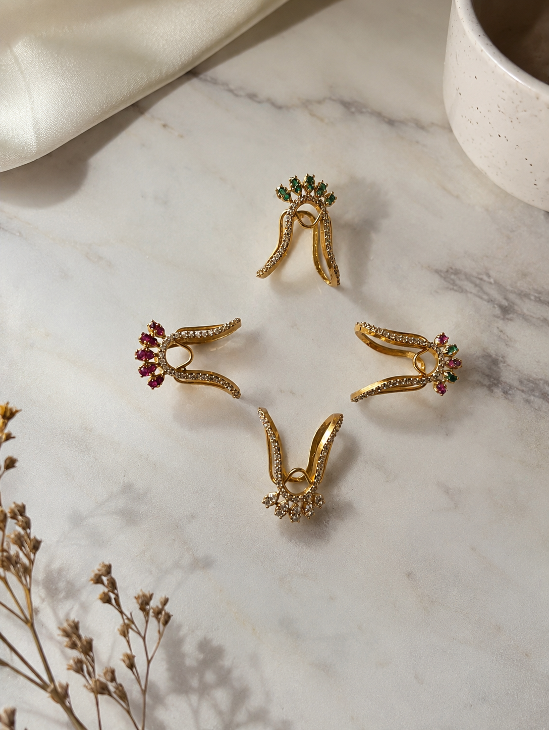 Four gold-toned Vanki chevron rings arranged in a diamond pattern on marble, showcasing the four color variants: emerald green, ruby red, clear white, and a dual-tone ruby-emerald combination.