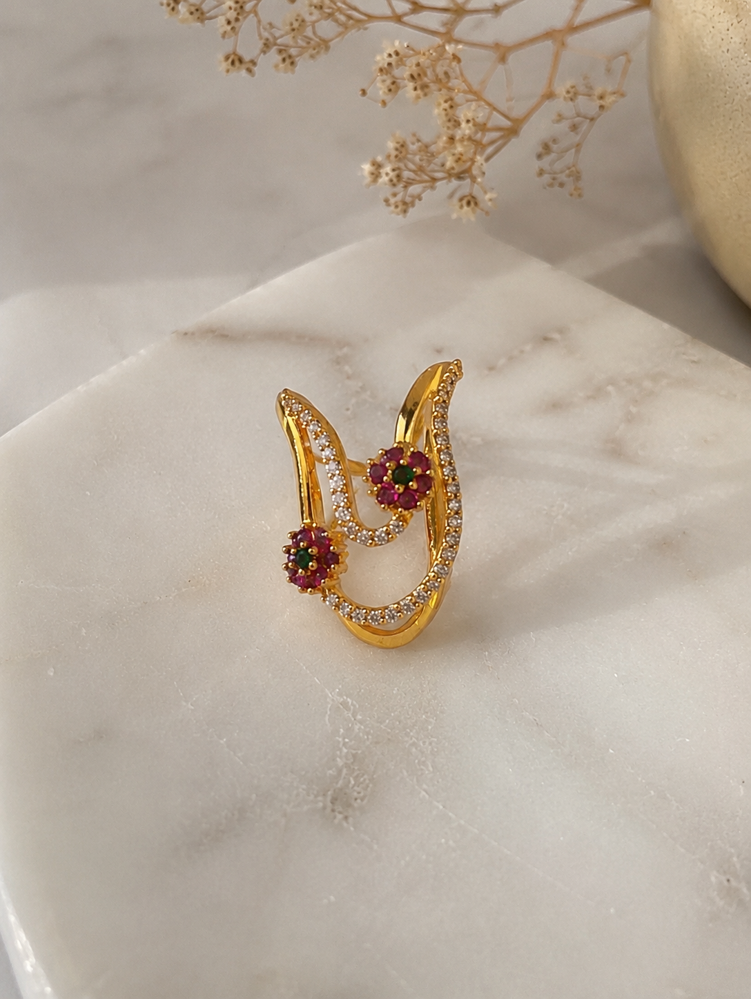 A gold-toned Vanki ring featuring pave American Diamond crystal bands and two floral clusters made of ruby-red and emerald-green stones on a marble surface.