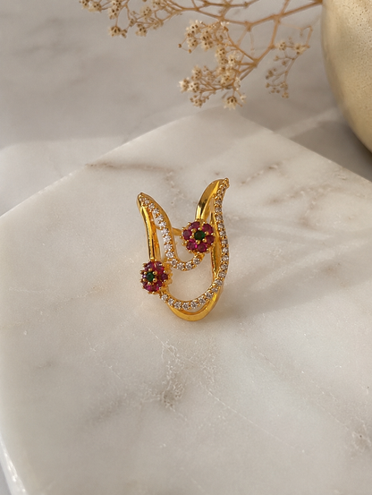 A gold-toned Vanki ring featuring pave American Diamond crystal bands and two floral clusters made of ruby-red and emerald-green stones on a marble surface.