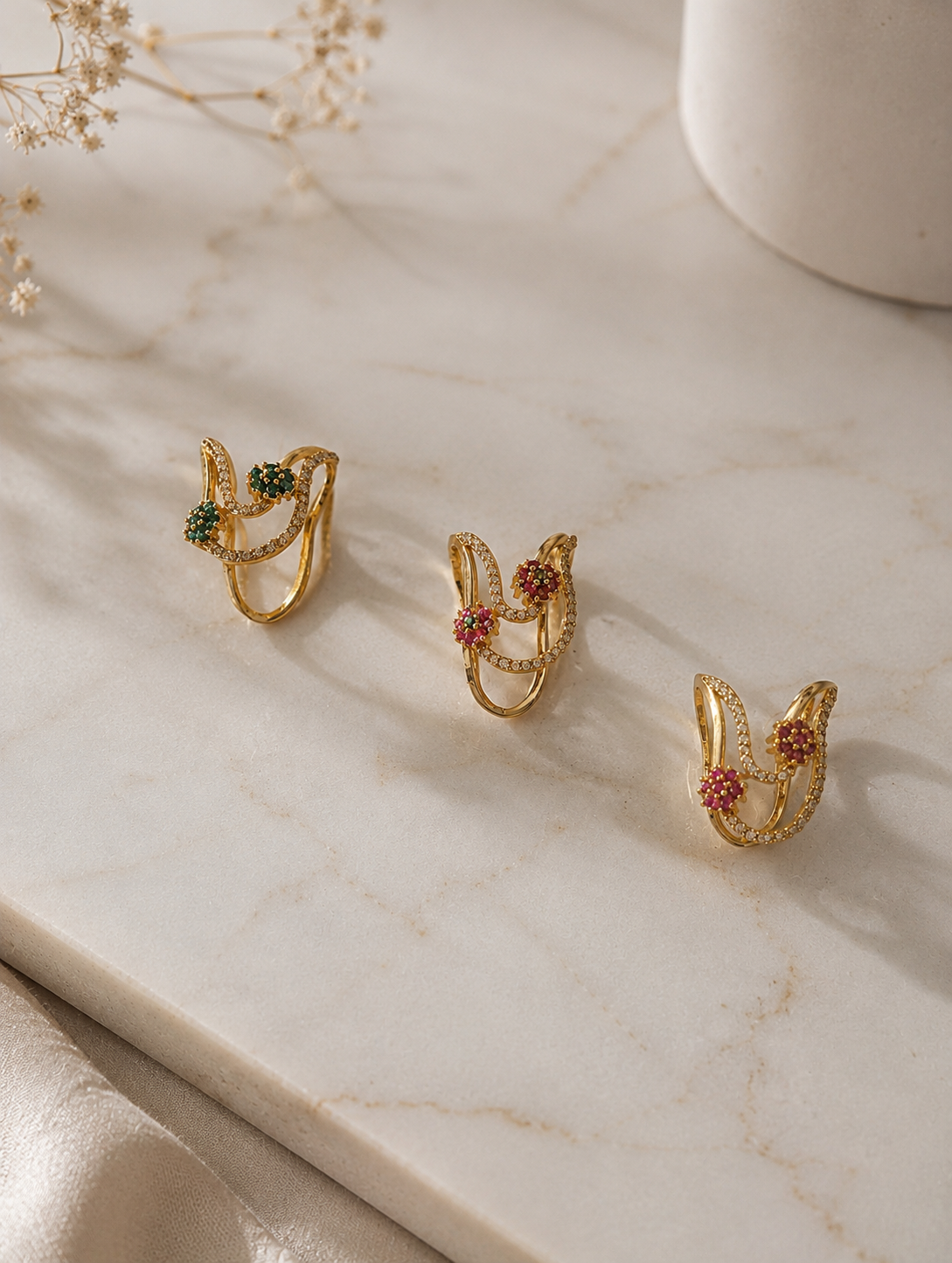 Three gold-toned floral Vanki rings arranged in a row on a marble slab, displaying emerald green, ruby pink, and dual-tone ruby-emerald stone variants.