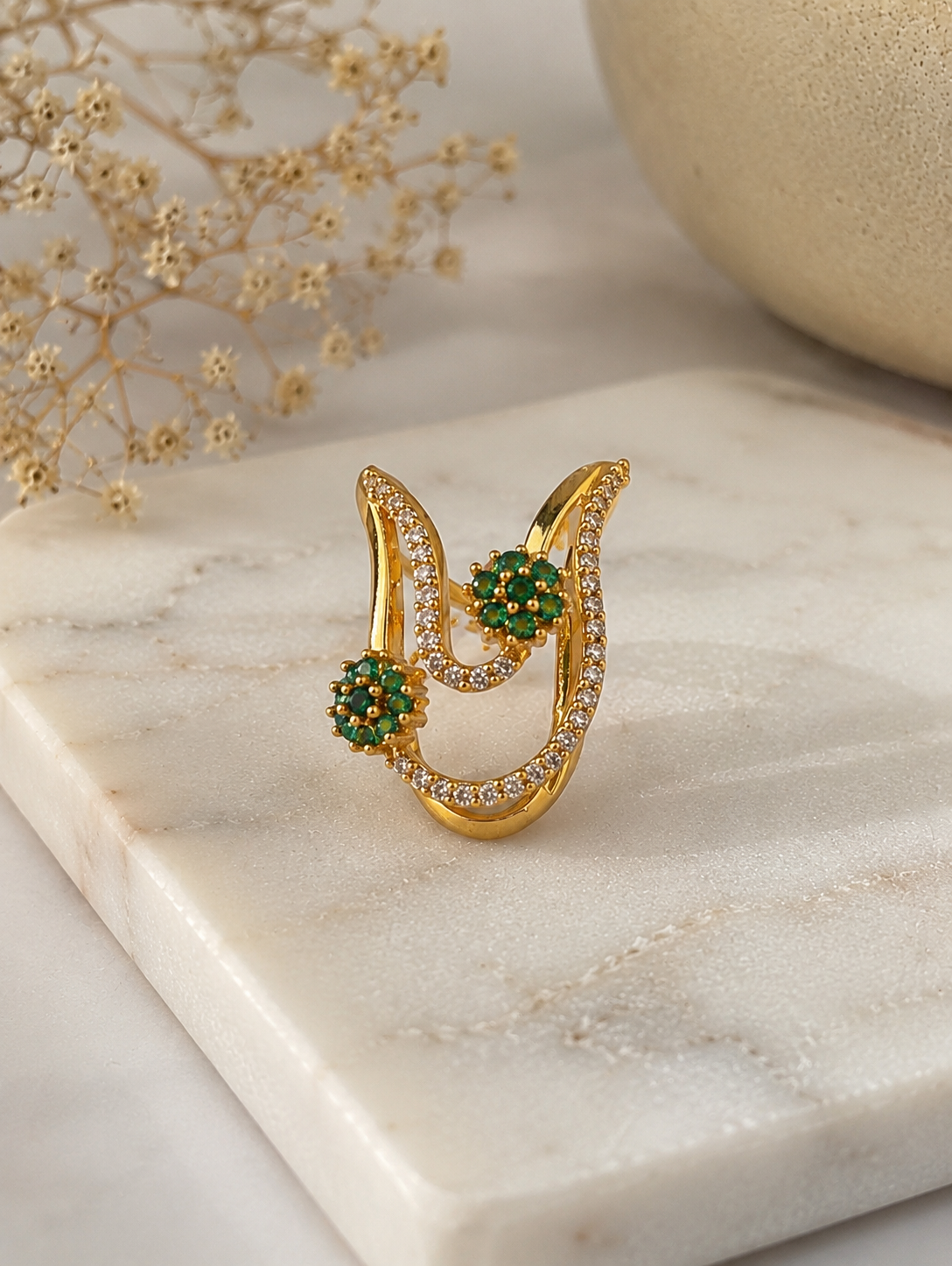 A close-up view of a gold-toned Vanki chevron ring adorned with pave clear CZ crystals and two emerald-green floral stone clusters resting on marble.