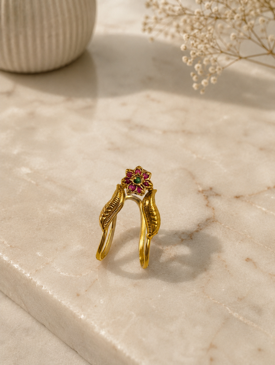 An antique gold-toned Vanki ring resting upright on a marble surface, featuring carved leaf details along its V-curved band and a central floral cluster encrusted with ruby-red stones and a green stone center.