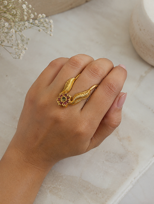 Close-up of a model wearing an antique gold-toned Vanki ring on her index finger, displaying leaf-engraved chevron arms topped with a ruby and green floral stone center motif.