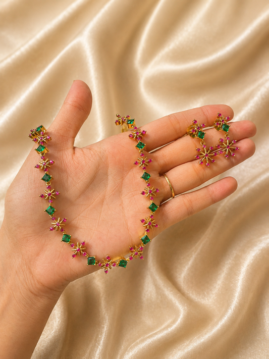 A person holding a gold-plated jewelry set featuring a delicate choker necklace and matching starburst drop earrings studded with square green emeralds and pink ruby stones against a champagne silk background.