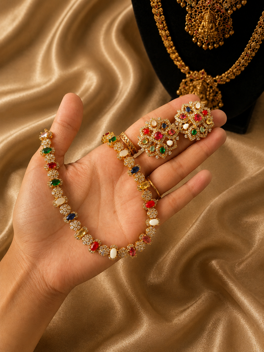A person holding a traditional gold-plated Navaratna jewelry set featuring a slender necklace and round floral stud earrings set with multi-colored gemstones and cubic zirconia diamonds against a gold silk background.