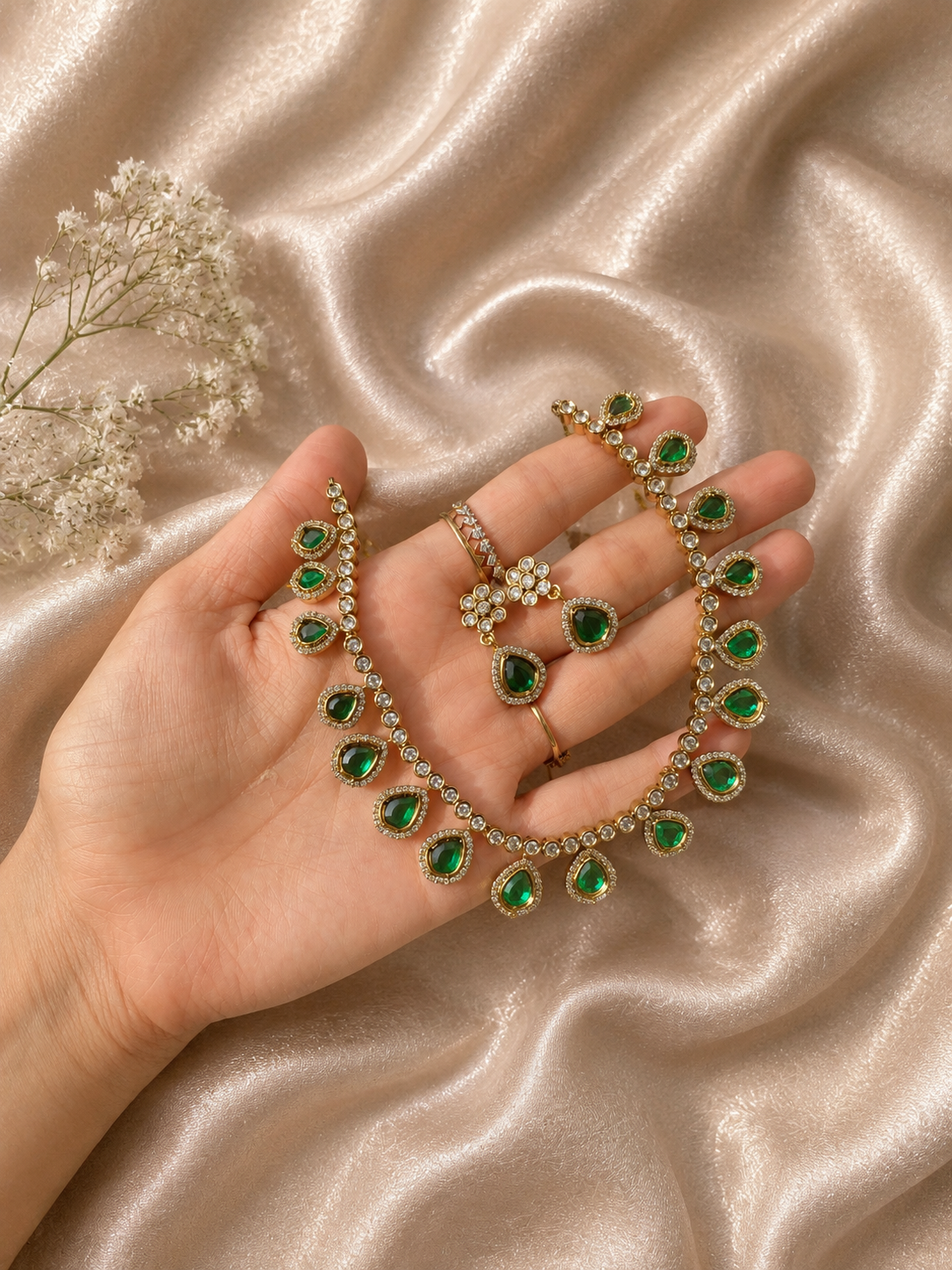 A person holding a delicate gold-plated jewelry set featuring a choker necklace and matching drop earrings designed with teardrop-shaped emerald green stones bordered by tiny white cubic zirconia crystals, set against a shimmering champagne silk fabric.