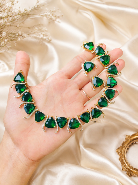 A person holding a gold-plated fashion jewelry set featuring a collar necklace and matching earrings made of large, vibrant, trilliant-cut emerald green gemstones bordered by sparkling white CZ crystal curves, displayed on a gold silk fabric.