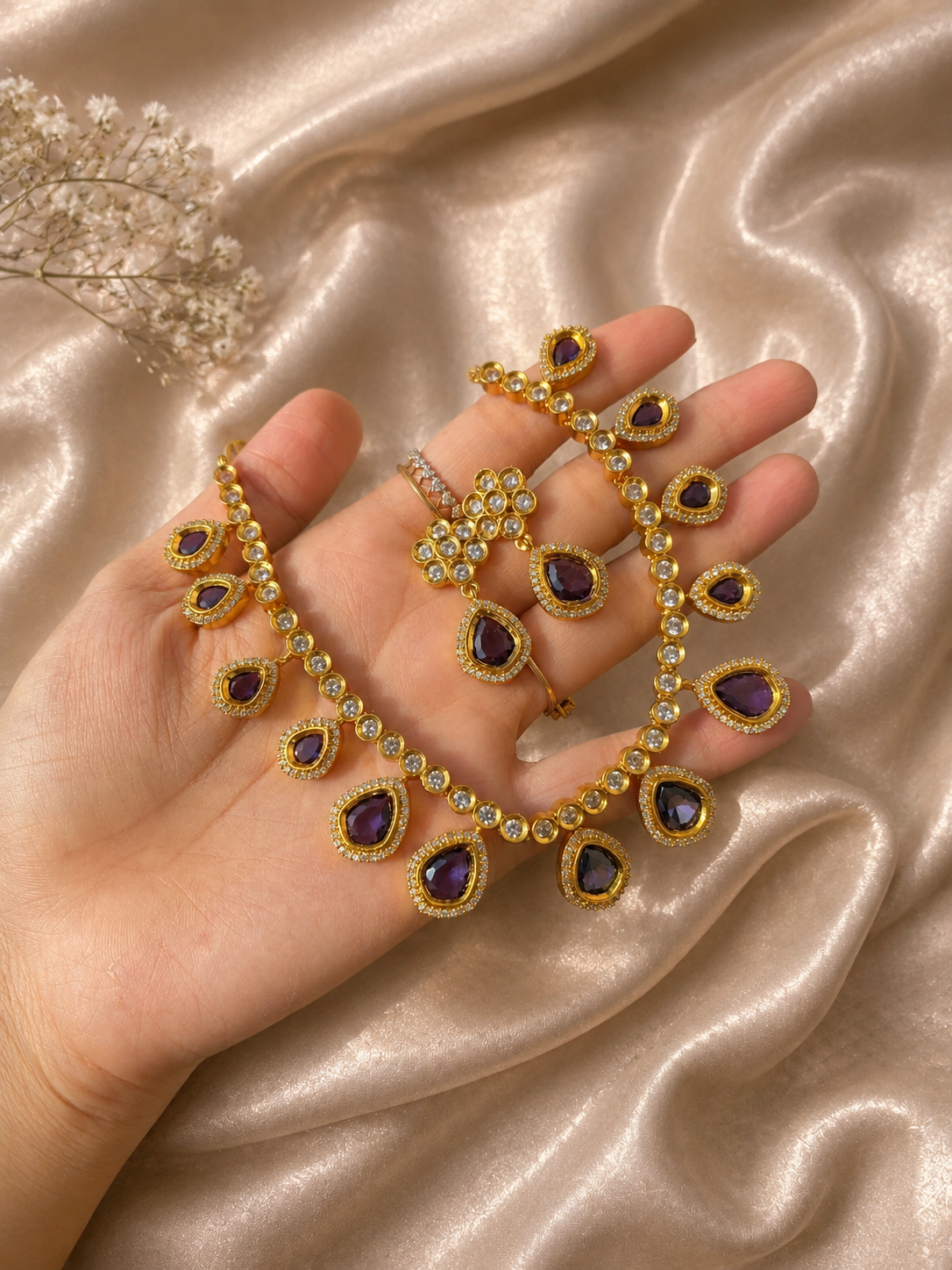 A person holding a delicate gold-plated jewelry set featuring a choker necklace and matching drop earrings designed with teardrop-shaped purple amethyst stones bordered by tiny white cubic zirconia crystals, set against a shimmering champagne silk fabric.