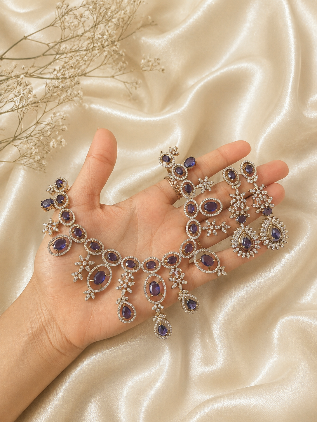 A person holding an elaborate silver-plated bridal jewelry set featuring a wide choker necklace and long matching drop earrings studded with oval and teardrop purple stones amidst sparkling white CZ crystal clusters, displayed against a cream silk background.