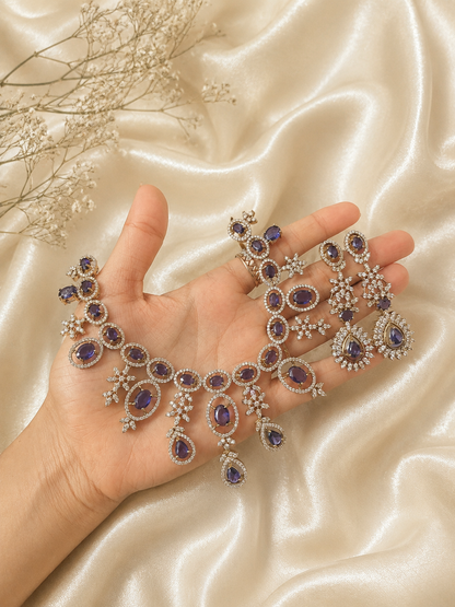A person holding an elaborate silver-plated bridal jewelry set featuring a wide choker necklace and long matching drop earrings studded with oval and teardrop purple stones amidst sparkling white CZ crystal clusters, displayed against a cream silk background.