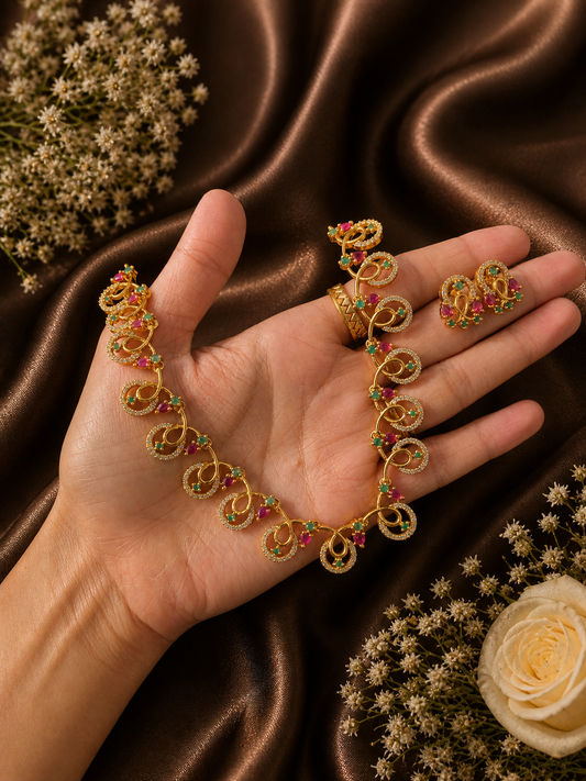 A person holding a gold-plated jewelry set featuring a necklace with a delicate wave and swirl design, studded with tiny pink rubies, green emeralds, and white CZ crystals, complete with matching small stud earrings against a brown satin fabric background.